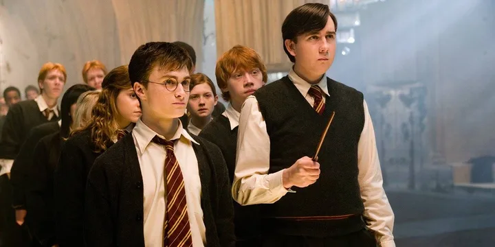 Harry Potter and Neville Longbottom in Hogwarts uniforms holding a wand with students behind them.