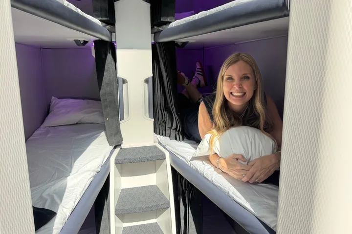 Smiling woman lying on an Air New Zealand Skynest bunk bed pod, holding a pillow, with purple-lit sleeping berths visibl