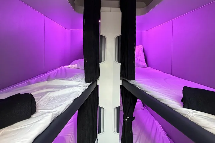 Air New Zealand Skynest bunk bed pods with white bedding and dark privacy curtains under vivid purple cabin lighting.