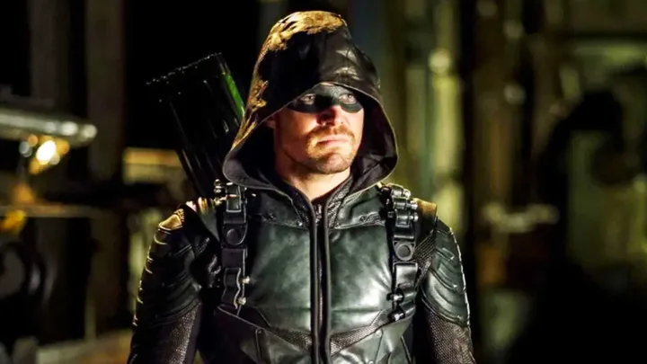 Male actor in green Arrow superhero costume with hood and mask in dark industrial setting.